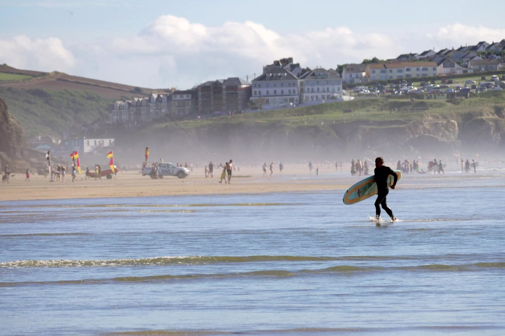 Perranporth beach looking towards the town, with lifeguard flags on the sand and a surfer walking out of the shallows